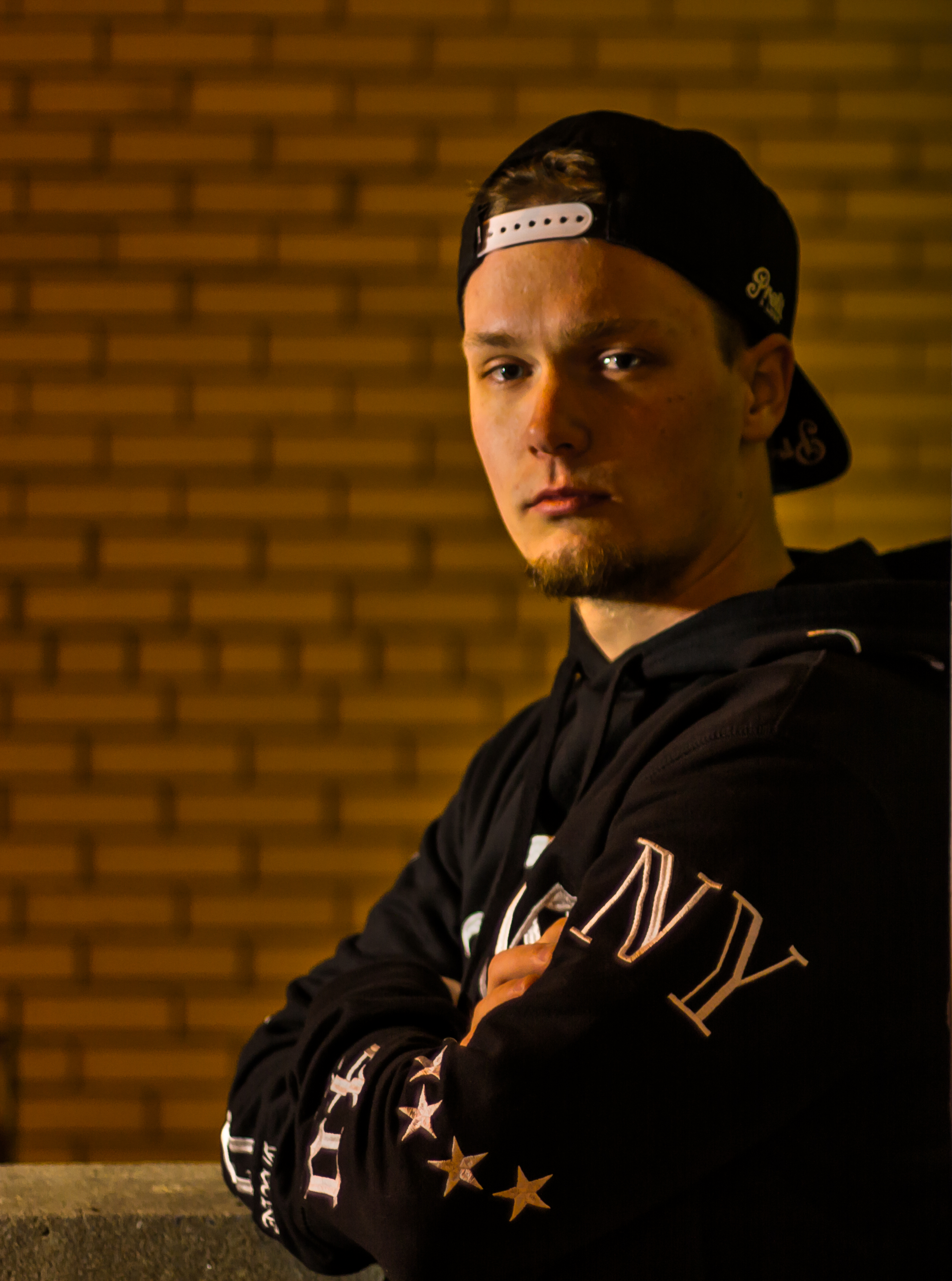 Artist portrait in front of a warm brick wall, wearing a backwards black cap and black jacket with arms crossed.